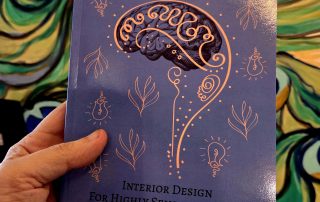 Hand holding the of the book, The Eccentric Genius Habitat Intervention, with a brain on the cover and a peacock mural behind it