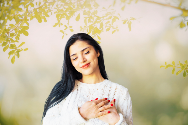 Trust Your Body Woman smiling, eyes closed, hands clasped over heart, dappled light and leaves in background